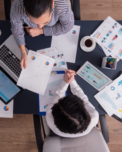 Top view of office workers meeting analyzing financial charts holding clipboard. Manager and employer discussing marketing strategy during briefing. Pointing at graphs.